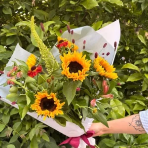 Bouquet champêtre artisanal composé de tournesols jaunes, de feuillage vert et de fleurs bordeaux, enveloppé dans du papier blanc avec un ruban pourpre. Création florale d'été.