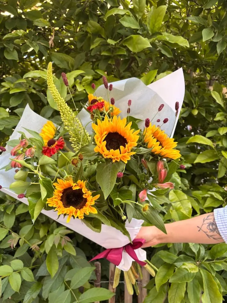 Bouquet champêtre artisanal composé de tournesols jaunes, de feuillage vert et de fleurs bordeaux, enveloppé dans du papier blanc avec un ruban pourpre. Création florale d'été.